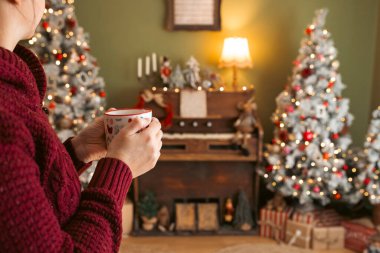 Işıklı Noel ağacının arka planında sıcak bir fincan kahveyi tutan eller. Sıcak bir ev, atmosferik kış ikilisi.. 