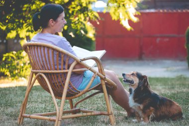 Kadın kitap okuyor, güneşli bir günde tüylü kahverengi köpeğiyle sandalyede dinleniyor. Hayvanlarla bağ kurma ve birlikte olmaktan keyif alma kavramı.