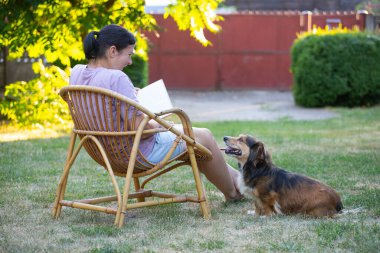 Kadın kitap okuyor, güneşli bir günde tüylü kahverengi köpeğiyle sandalyede dinleniyor. Hayvanlarla bağ kurma ve birlikte olmaktan keyif alma kavramı.