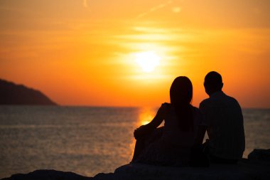 Çift kayalarda oturuyor ve denizin üstünde turuncu günbatımının tadını çıkarıyor..