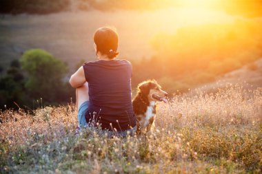 Rahatlamış kadın ve köpek tepenin üstünde yaz günbatımının tadını çıkarıyorlar..