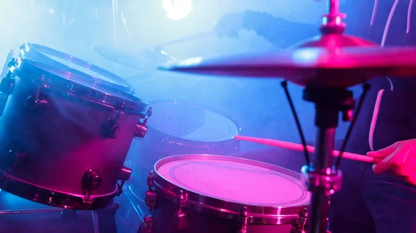 A drummer plays on a dark stage in the fog and neon lights.