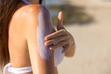 Bikinili genç bir kadın yazın plajda güneş kremi sürüyor.