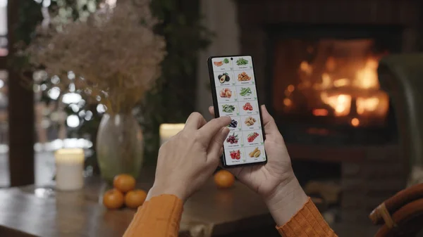 在家里用智能手机点菜 一个女人用智能手机上的应用程序在网上商店里挑选蔬菜 炉火熊熊燃烧的家居装饰 图库图片