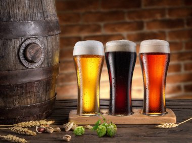 Three chilled glasses of different beer and beer barrel on wooden table closeup. 
