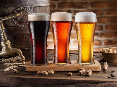 Three chilled glasses of different beer and beer barrel on wooden table closeup. 