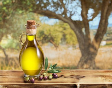 Bir şişe zeytinyağı ahşap bir masanın üzerinde duruyor. Arkasında da biraz bokeh, güneşli bir günde zeytin bahçesi var..