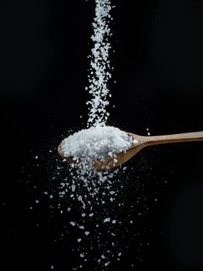 Siyah arka planda yenilebilir tuz kristalleri tahta kaşığa dökülüyor..