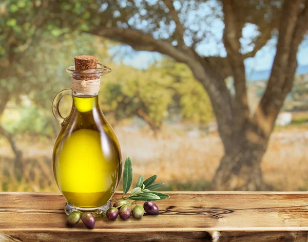 木桌上立着一壶手工种植的橄榄油 后面是一个略显潮湿 阳光灿烂的橄榄园 — 图库照片