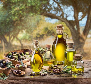 Bir şişe zeytinyağı ahşap bir masanın üzerinde duruyor. Arkasında da biraz bokeh, güneşli bir günde zeytin bahçesi var..