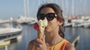 Güneş gözlüklü genç kız dondurma külahı yiyor. Arka planda, Bokeh ve yatları deniz koyunda..