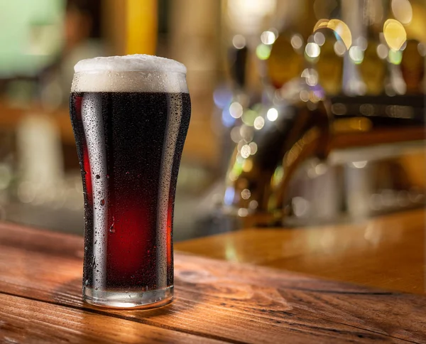 Glass of chilled stout beer on wooden bar table top and blurred bar interior at the background.