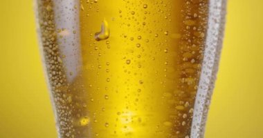 Kamera bira bardağı boyunca hafif birayla yavaşça yükselir. Bardakta bir sürü buhar damlası ve üstünde bira köpüğü var. Sarı arkaplan. 