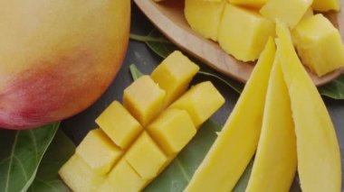 Olgun mango meyveleri, dilimler ve mango yapraklarıyla gri taş bir masanın üzerinde yavaşça hareket eder. Projelerin için güzel bir meyve arka planı. Makro video çekimi.
