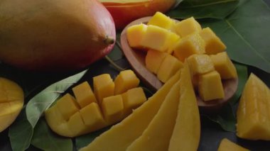 Olgun mango meyveleri, dilimler ve mango yapraklarıyla gri taş bir masanın üzerinde yavaşça hareket eder. Projelerin için güzel bir meyve arka planı. Makro video çekimi.