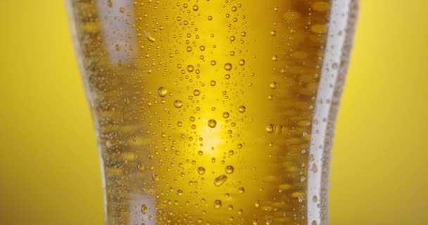 カメラは軽いビールでビールグラスに沿ってゆっくりと上昇します ガラスに凝縮ドロップがたくさんあり ビールの泡が上にあります イエローバック — ストック動画