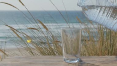 Su plastik bir şişeden bardağa dökülüyor, arka planda okyanus manzarası. Yavaş çekim.