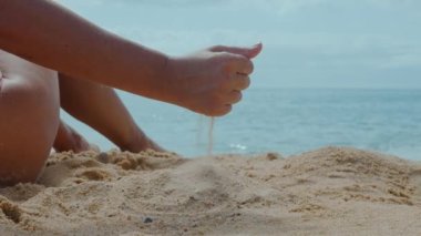 Kadın güneşli bir yaz gününde kumlu bir sahilde oturur ve elinden küçük bir kaydırağın üzerine kum döker. Bacakları çerçevede görünüyor. Sinema stili.