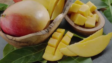 Olgun mango meyveleri, dilimler ve mango yapraklarıyla gri taş bir masanın üzerinde yavaşça hareket eder. Projelerin için güzel bir meyve arka planı. Makro video çekimi.
