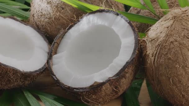 ココナッツスライスとココナッツの葉とともに新鮮なオープンココナッツが木のテーブルにゆっくりと回転しています あなたのプロジェクトのための素晴らしいフルーツの背景 ココナッツミルクはココナッツハーフの1つに注ぎ込まれています — ストック動画