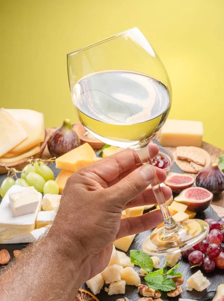 Glass of white wine in a man\'s hand with variety of sliced cheeses with fruits, mint and nuts. Yellow background. Wonderful wine background for your projects.