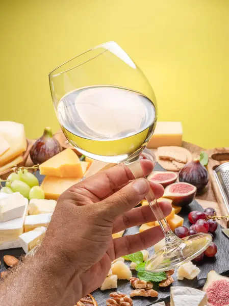 Glass of white wine in a man\'s hand with variety of sliced cheeses with fruits, mint and nuts. Yellow background. Wonderful wine background for your projects.
