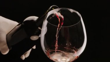 Kırmızı şarap siyah arka plandaki bir bardağa dökülüyor. Yavaş çekim, yakın çekim. Projeleriniz için harika bir şarap geçmişi var..