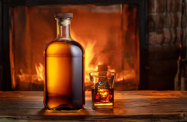 Glass of whiskey and whiskey bottle on old wooden table and blurred fireplace at the background.