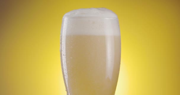 ビールのグラスは黄色の背景でゆっくり回転し ビールグラスには凝縮があり 中には泡や泡がたくさんあります あなたのプロジェクトのための素晴らしいビールの背景 — ストック動画