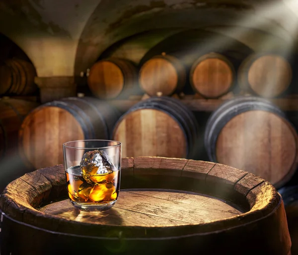 Glass of whiskey on top of  wooden barrel and  wood barrels in cellar at the background. Sun ray falling down on it surface. Placement for your product.