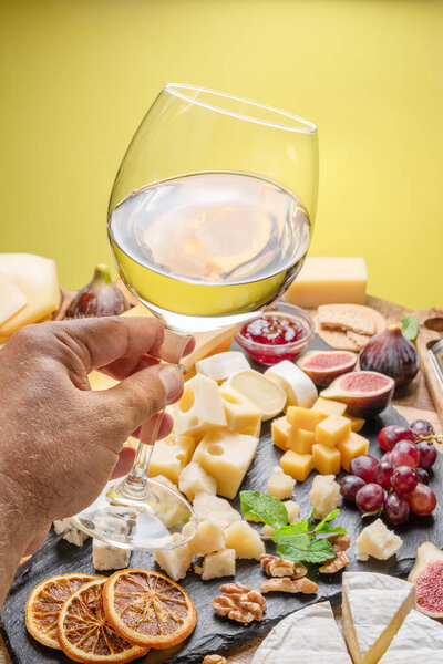 Glass of white wine in a man's hand with variety of sliced cheeses with fruits, mint and nuts. Yellow background. Wonderful wine background for your projects.