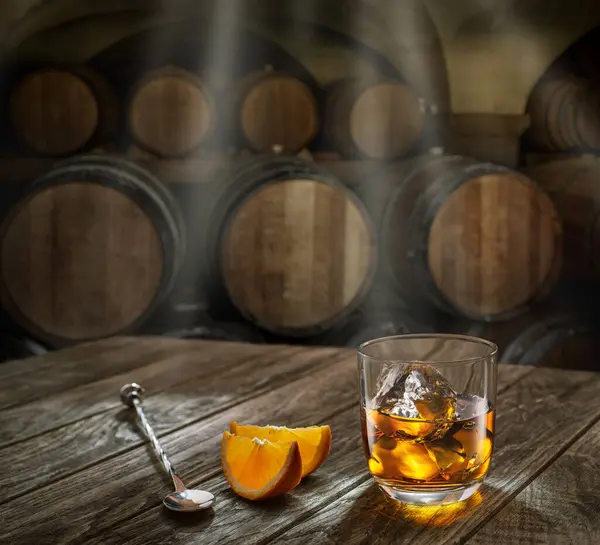 Glass of whiskey on top of  wooden barrel and  wood barrels in cellar at the background. Sun ray falling down on it surface. Placement for your product.