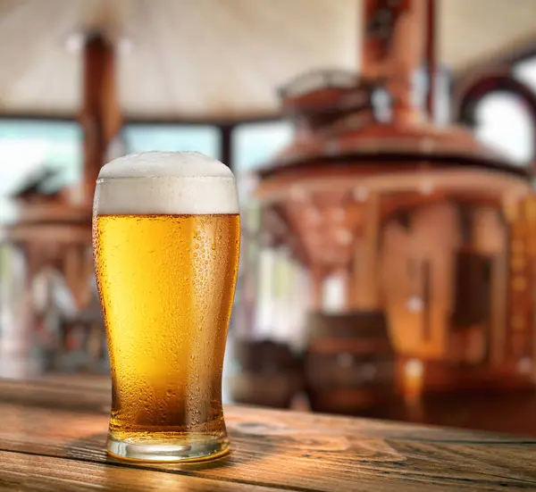 木制桌子上的冰镇淡啤酒和背景上模糊的铜酿造系统 图库图片