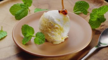 Karamel şurubu beyaz-sarı bir top dondurmanın üzerine dökülür; ayrıca tabakta taze nane yaprakları vardır. Projeleriniz için harika bir dondurma geçmişi var..