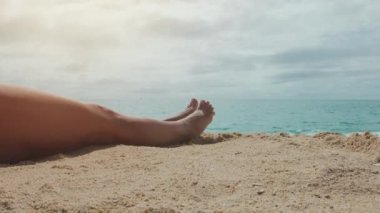 Güneşli bir yaz gününde kumlu bir sahilde oturan bir kadın. Ön planda güzel dişi bacakları yavaşça hareket ediyor. Sinema stili.