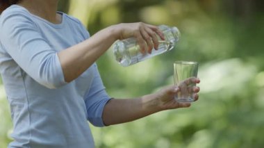 Kadın şişeden bir bardağa saf su döküyor, bulanık yeşil bir arka plan..