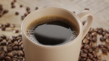 Kahve çekirdekli ahşap bir masanın üzerinde beyaz bir fincan kahve ve yükselen buhar yavaşça döner. Projeleriniz için güzel bir sıcak kahve arka planı..