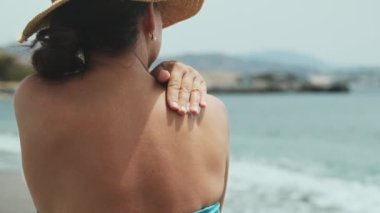 Sahilde otururken omzuna güneş kremi süren bir kadın. Deriyle ilgilenmek ve deriyi güneşten korumak. Sağlıklı tatil kavramı.