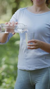 Kadın şişeden bir bardağa saf su döküyor, bulanık yeşil bir arka plan..