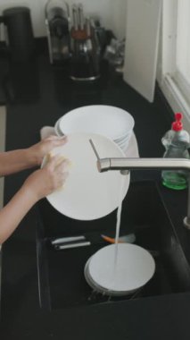 Kadın mutfak lavabosundaki bir su deresinin altında bulaşıkları elle yıkıyor ve duruluyor. Bulaşık yıkama işlemi. Yavaş çekim dikey video. 