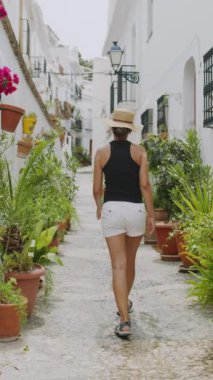 Beyaz şortlu ve güneş şapkalı orta yaşlı kadın İspanya 'da bir yerlerde uzun dar turistik sokaklarda yürüyor. Avrupa 'da tatil ve tatil kavramının yavaş çekim dikey görüntüleri. 