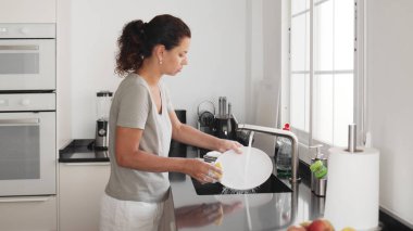 Kadın mutfak lavabosundaki bir su deresinin altında bulaşıkları elle yıkıyor ve duruluyor. Bulaşık yıkama işlemi.
