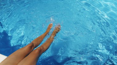 Woman is sitting at the edge of the pool with her legs dangling in the water and shaking the water  and making ripples and small waves. Summer background.  clipart