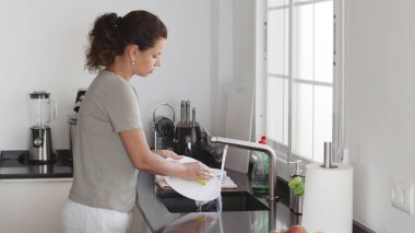 Woman washing and rinsing dishes by hand under a stream of water in the kitchen sink. Washing dishes process. clipart