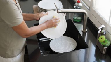 Woman washing and rinsing dishes by hand under a stream of water in the kitchen sink. Washing dishes process. clipart