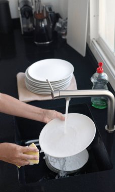 Washing and rinsing dishes by hand under a stream of water in the kitchen sink. Washing dishes process. clipart