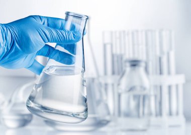 Mavi eldivenli konik matara ve arka planda laboratuvar ekipmanları..