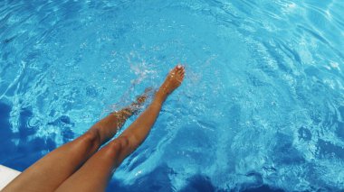 Woman is sitting at the edge of the pool with her legs dangling in the water and shaking the water  and making ripples and small waves. Summer background.  clipart