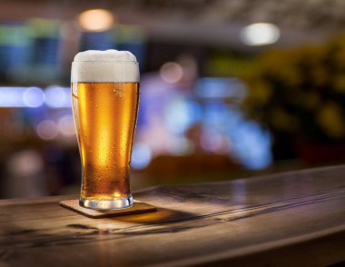 Bir bardak soğuk bira ahşap masa üstünde ve arka planda parlak ışıklı bar iç kısmı bulanık..