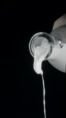 Siyah arka planda bir şişe sütten bardağa süt dökmek. Dikey yavaş çekim görüntüleri.